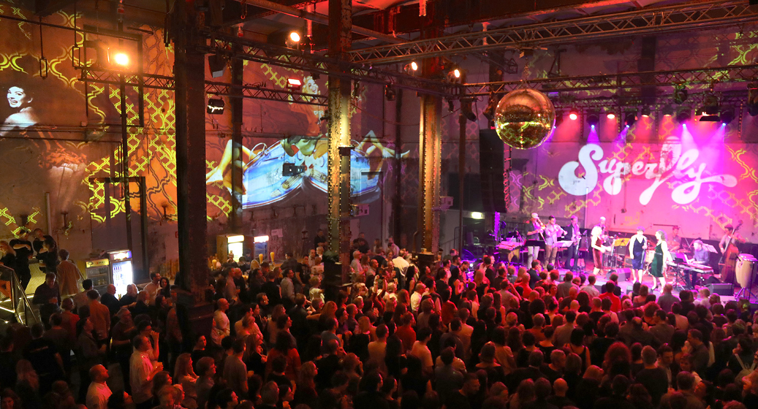 Band vor einer pinken Superfly-Projektion auf der Bühne – Superfly, Ottakringer Brauerei, Wien, Österreich